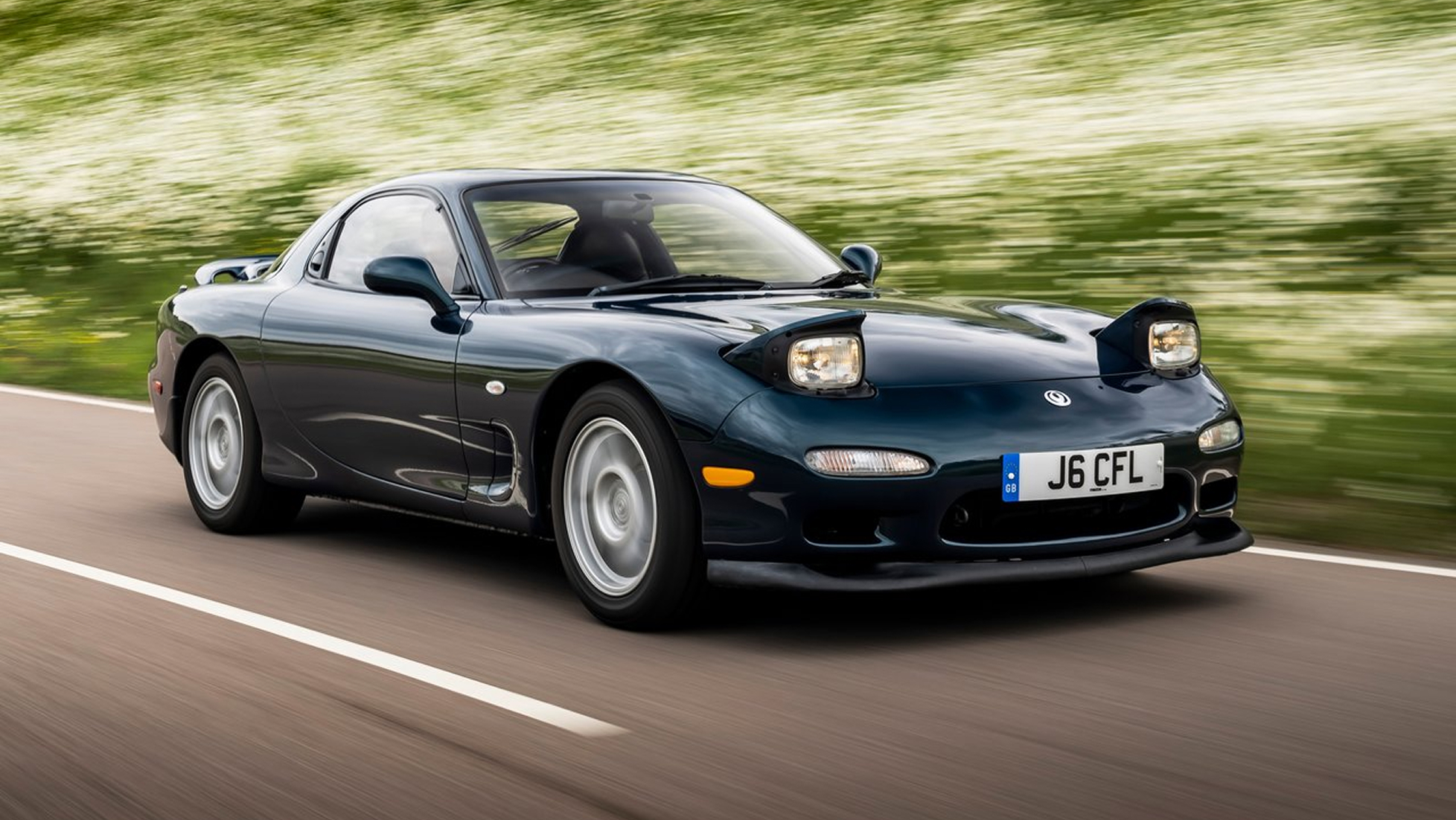 Black Mazda RX-7 FD driving on a country road with pop-up headlights raised